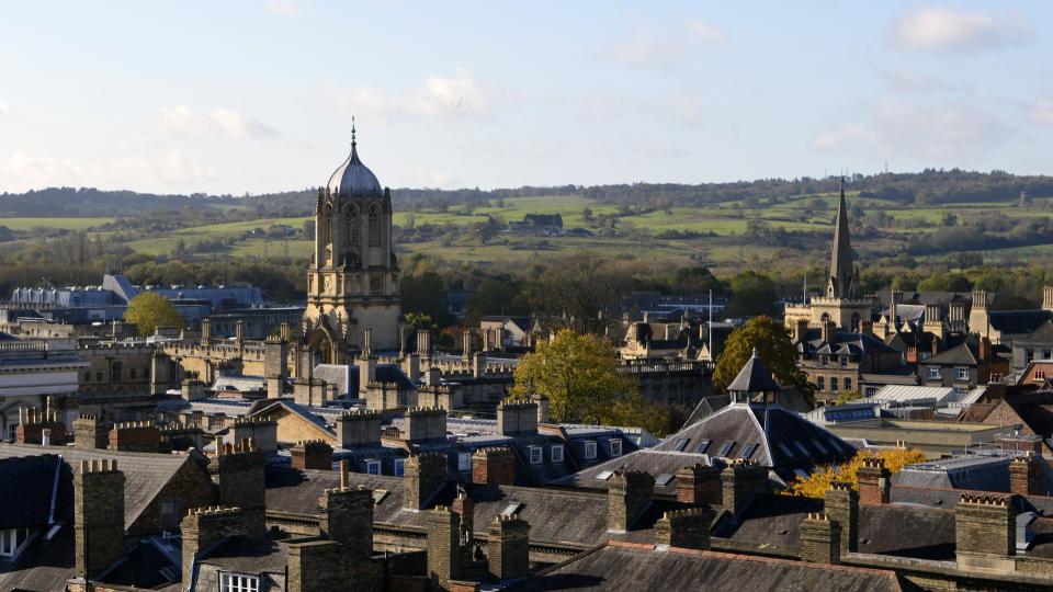 Photo overlooking the city of Oxford, showing fields and trees in the background and rooftops in the foreground.
