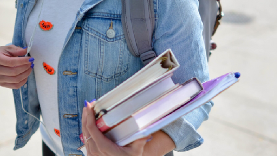 Student holding books and folders wearing a blue denim jacket and grey t-shirt.