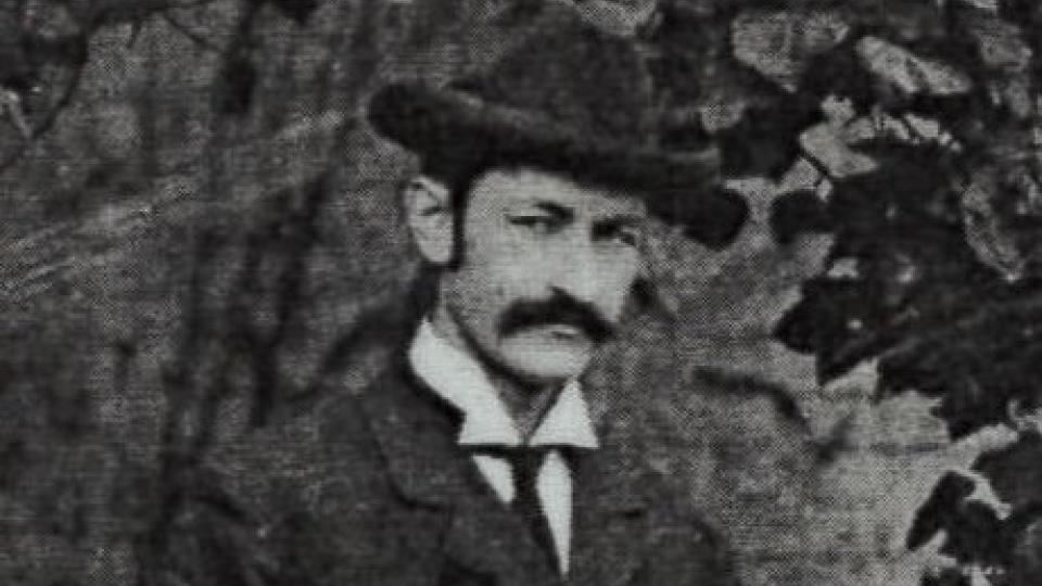 Walter Vrooman has a moustache and is wearing a suit, tie and dark hat.