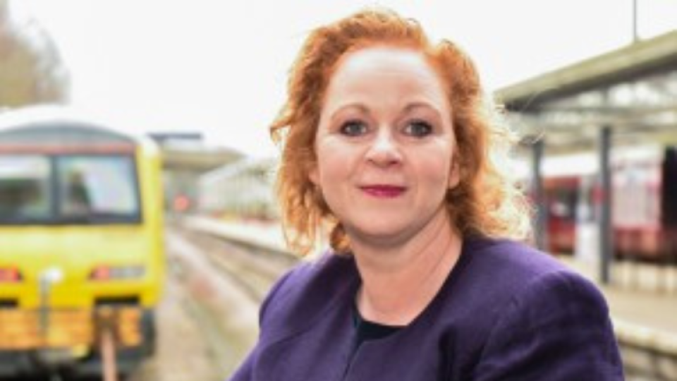 Judith Cummins has red hair and a purple suit on, she is standing at a railway station with a train behind her.