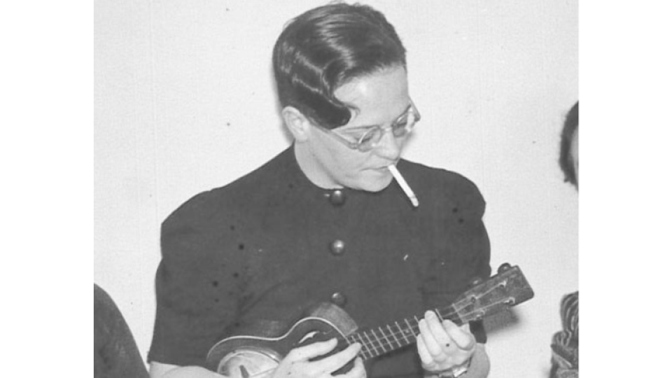 Kathleen McColgan has gelled hair, is smoking and wearing a long-sleeved shirt, whilst playing a ukulele.