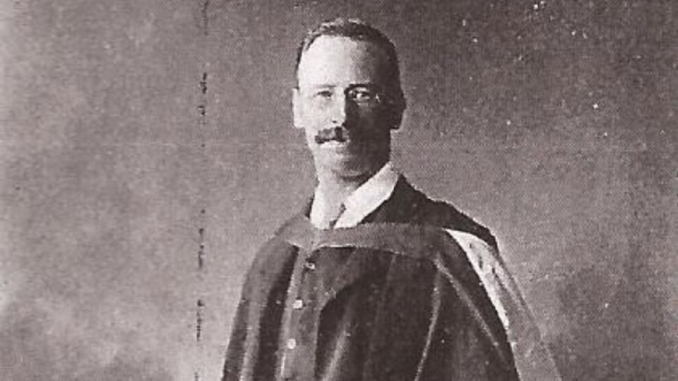 Gilbert Slater is wearing a gown. He has a moustache and glasses on.