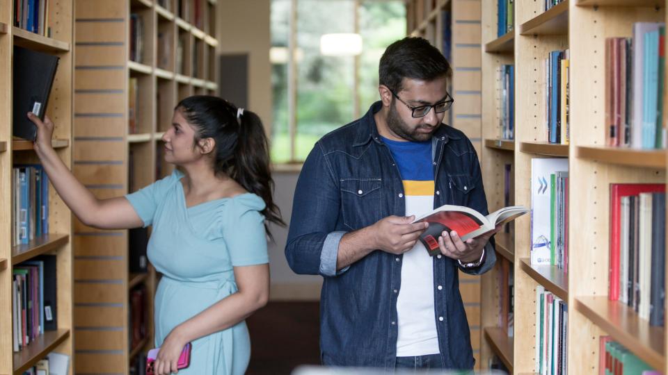 Two students in The Callaghan Library handling books, one reading, the other taking a book out of a book shelf.