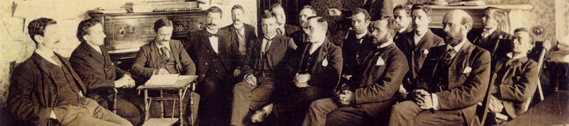 Early twentieth century photo of fifteen men in smart suits sat down facing each other.
